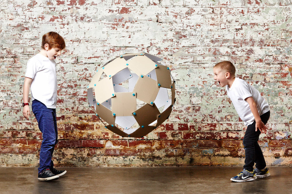 Two children toss an oversized ball between them. The ball is made from squares of cardboard connected using Makedo Scrus.